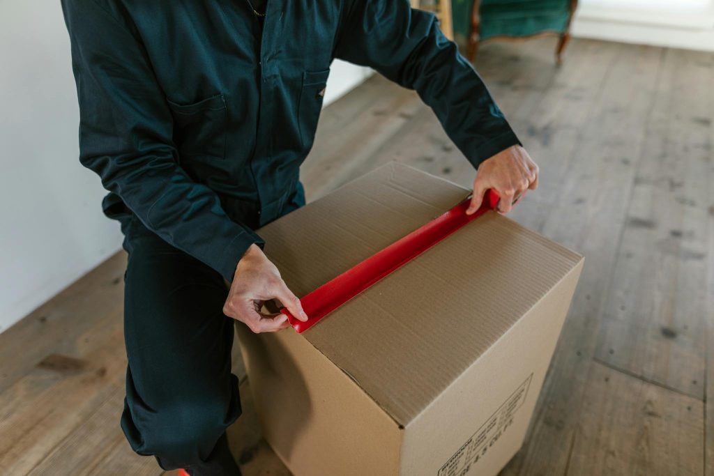 Male courier sealing a cardboard box with red tape, indoor wooden floor setting.